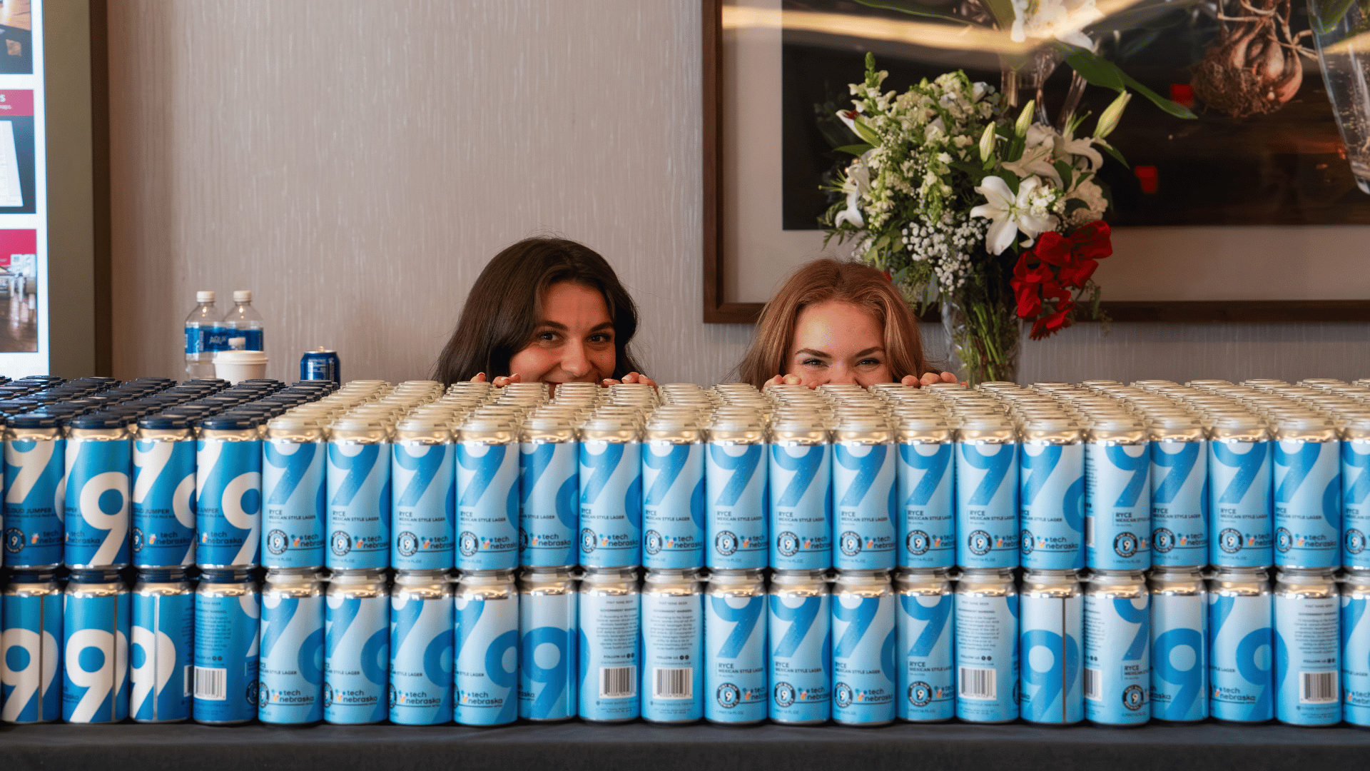 Two attendees peeking over a wall of Tech Nebraska branded cans