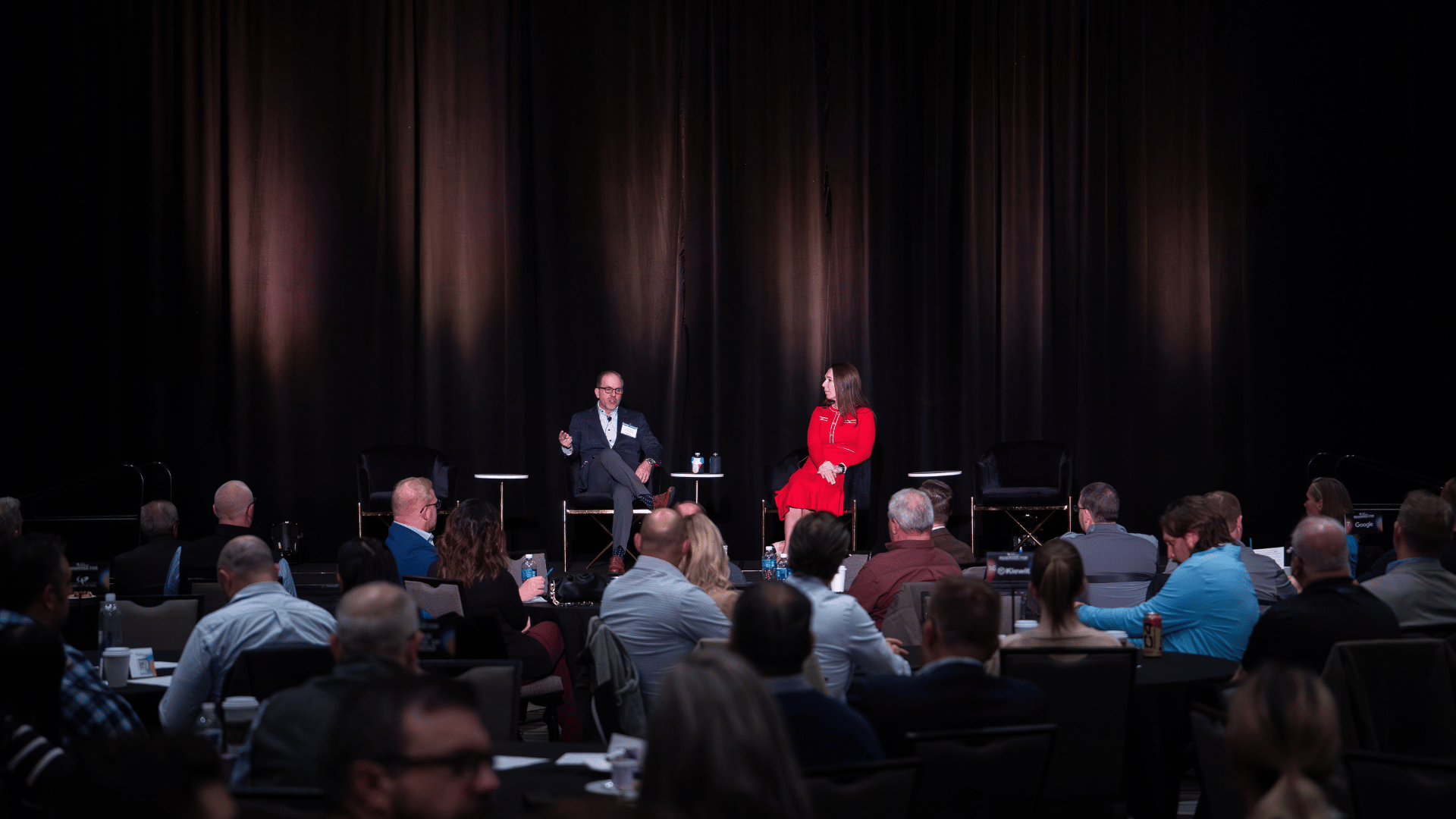 Fireside chat in front of a dark curtain with audience watching